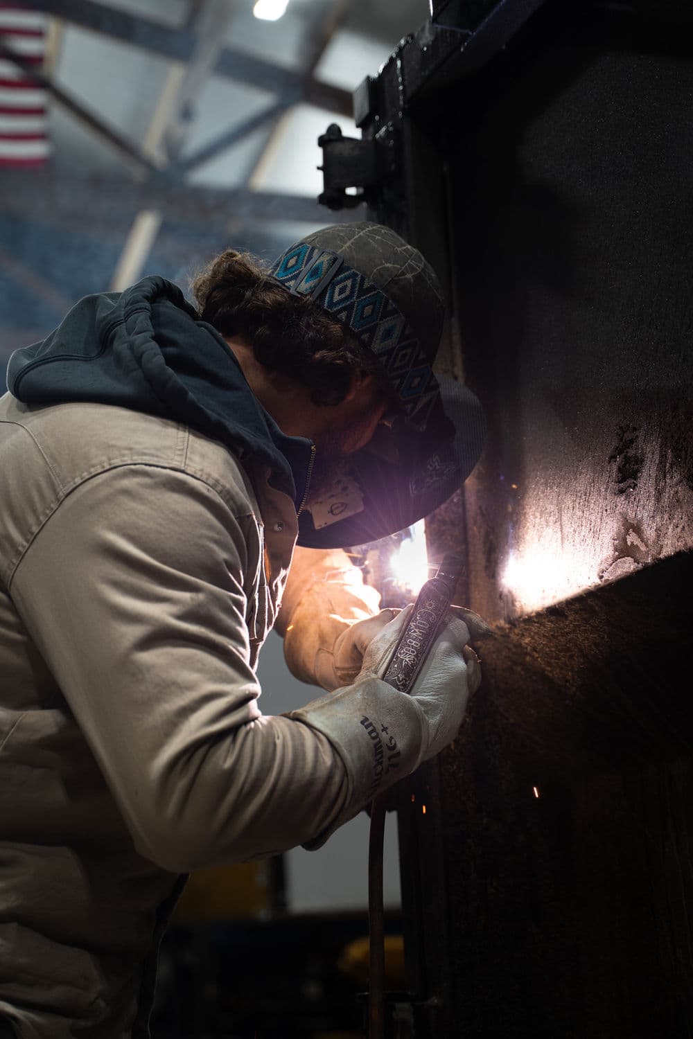 Welder in protective gear working on metal with sparks flying in industrial setting.