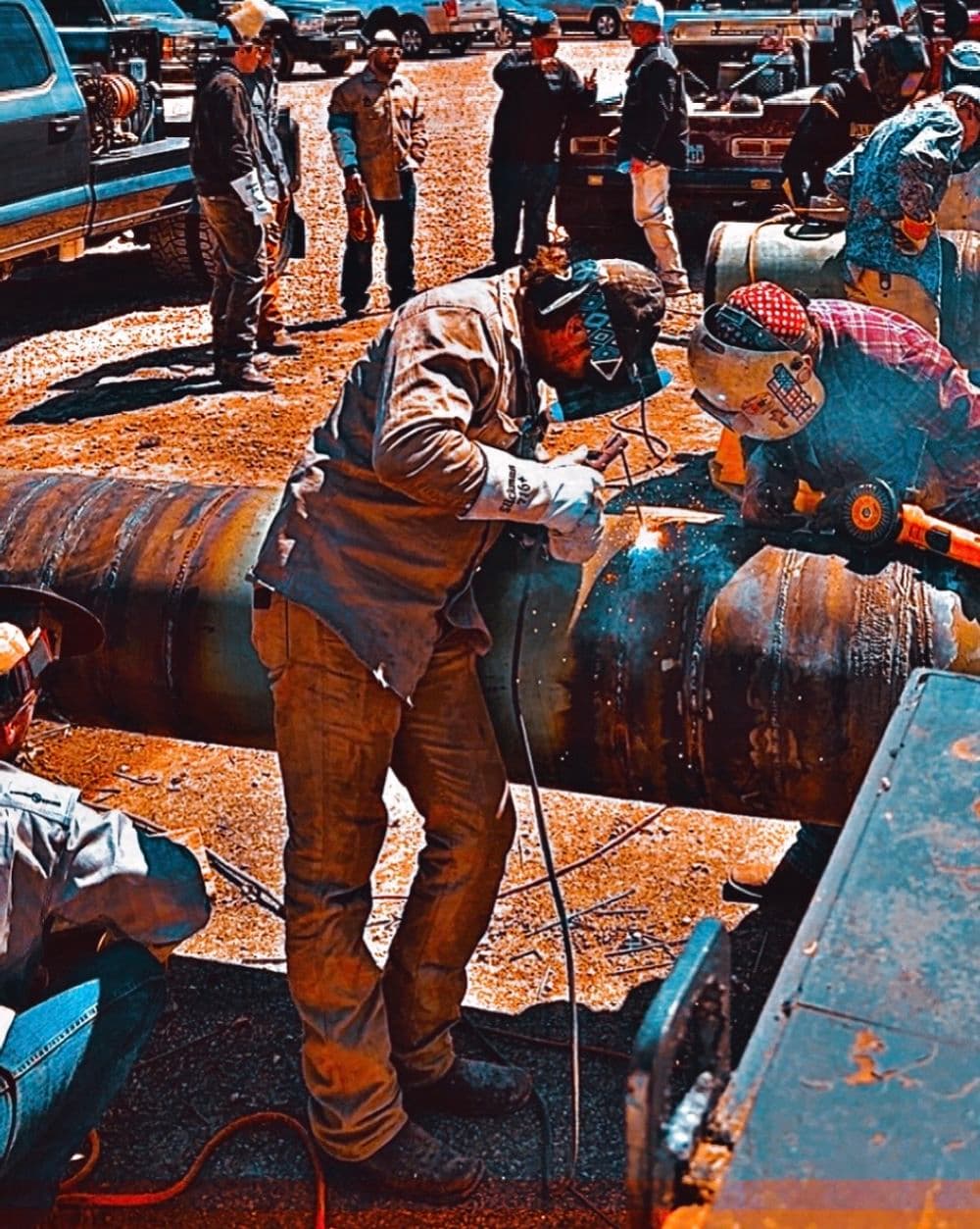 Welders working on pipes in an outdoor construction site with various vehicles in the background.